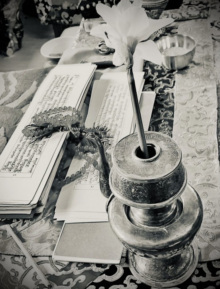 Buddhist shrine table with open Tibetan texts, a red mala, and a golden ritual vase, shown in black and white