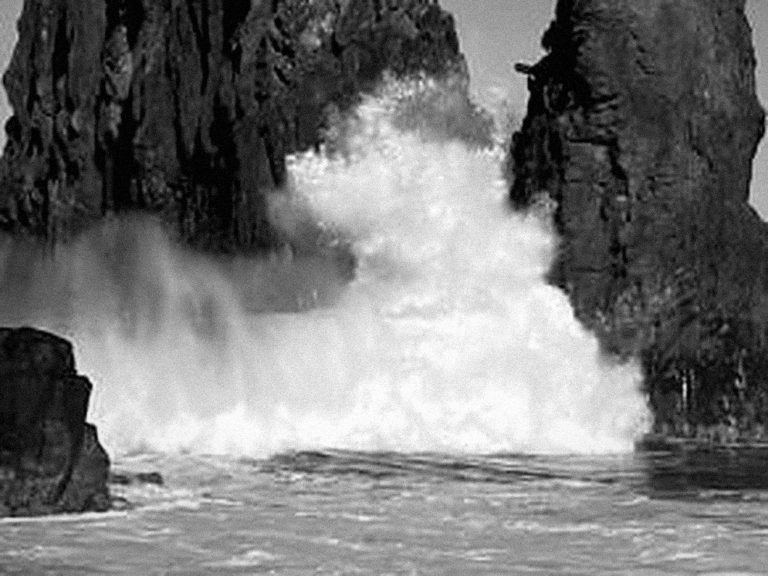 Black and white photo of an ocean wave breaking through rocky cliffs, spray rising into the air